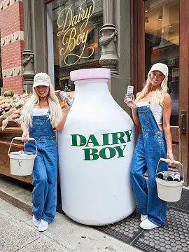 Two people in matching white tops, denim overalls, and caps pose on a city sidewalk holding metal pails beside a giant white milk-bottle prop labeled “DAIRY BOY” outside a storefront.