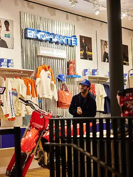 Interior of a trendy retail store with a neon “ENCHANTÉ” sign, colorful apparel and hats on display, and a person in a cap smiling beside a bright red motorcycle.