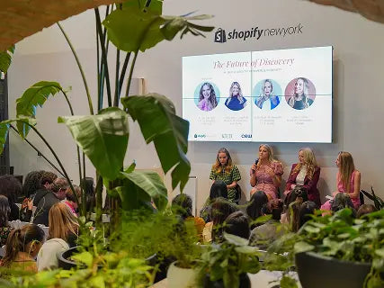 Panel discussion at a Shopify New York event, with four speakers seated in front of an audience and a large screen behind them titled “The Future of Discovery,” surrounded by lush indoor plants.