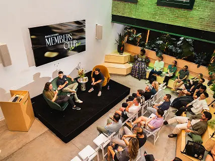 Audience seated in a modern event space watching a live conversation between two speakers on a small stage, with a large screen reading “Members Club” behind them and greenery along the walls.