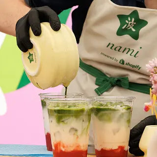 Close-up of a barista wearing a “nami powered by Shopify” apron, pouring green matcha from a bowl into clear cups layered with milk and red fruit at the bottom.