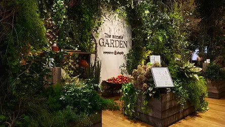 Immersive indoor garden installation with dense green foliage, flowers, and produce displays surrounding a white wall sign that reads “The Sonsie GARDEN powered by Shopify.”